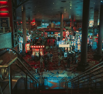 A bustling casino floor filled with groups of slot machines and people walking around. The atmosphere is vibrant with neon lights and illuminated signs. The interior has a ceiling supported by large pillars and there are stairs leading down to the gaming area.